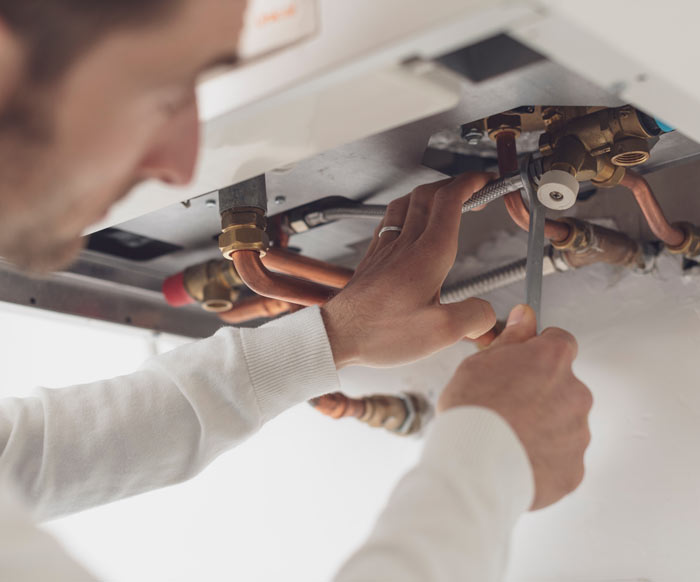 A person performing plumbing work, adjusting pipes and valves under a sink with copper and metal fittings.