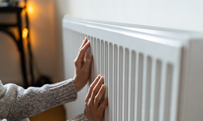 A person gently touching a white radiator with their hands, wearing a cozy sweater, in a softly lit room.