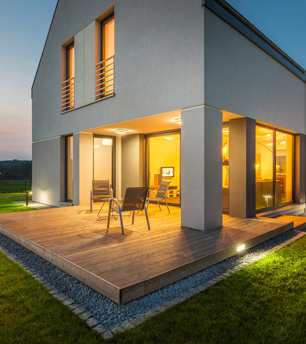 Modern house exterior with a wooden deck, featuring two chairs, illuminated by warm light at dusk, surrounded by green grass and pebbles. Ideal for outdoor tranquility and relaxation.