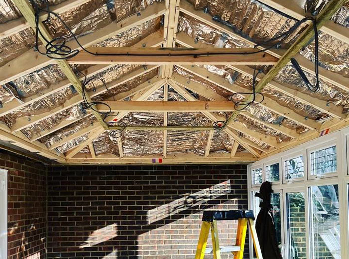 Interior view of a room under construction, featuring a wooden ceiling framework with silver insulation material, electrical wiring, and a step ladder. Home renovation progress is visible, highlighting structural elements.