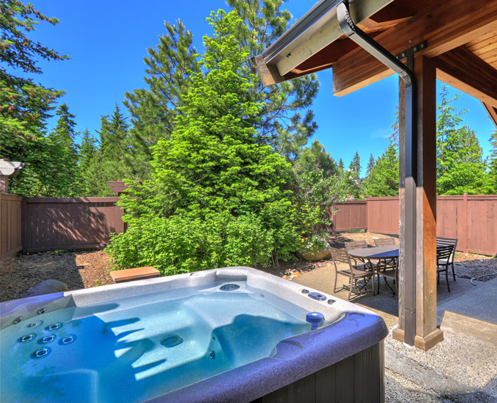 A view of a hot tub in a serene outdoor setting, surrounded by lush green trees and a patio area with a table and chairs under a wooden structure, ideal for relaxing in a peaceful retreat.