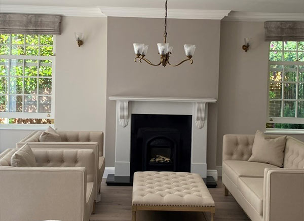 Stylish living room featuring beige upholstered sofas, a tufted ottoman, a classic fireplace, and elegant light fixtures, creating a cozy and inviting atmosphere.