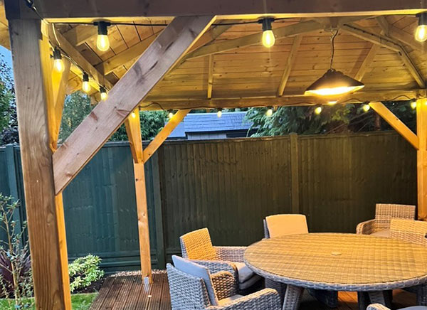 A cozy outdoor gazebo with string lights, featuring a round table surrounded by wicker chairs, set against a green wooden fence, perfect for gatherings.