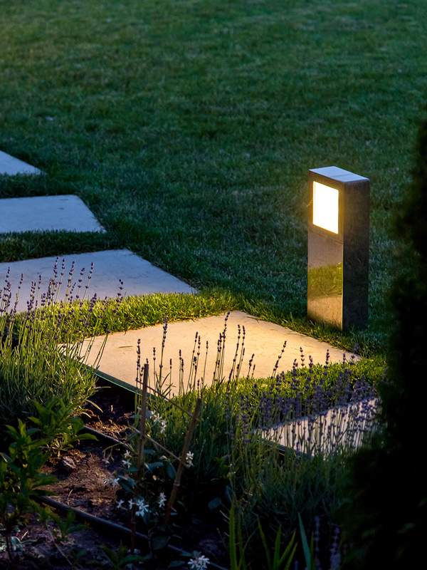 Outdoor pathway illuminated by a square light fixture, surrounded by lush green grass and lavender plants at dusk. Pathway lighting enhances the garden's aesthetics.