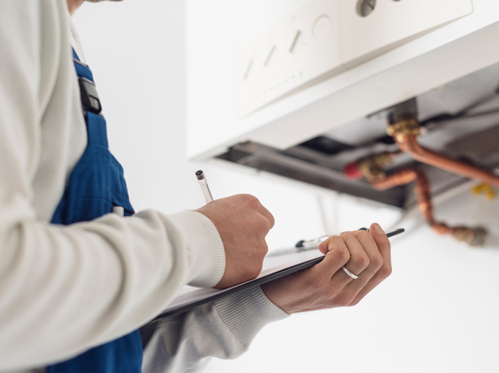 A technician in blue overalls writing notes while inspecting a heating system, including pipes and controls above.