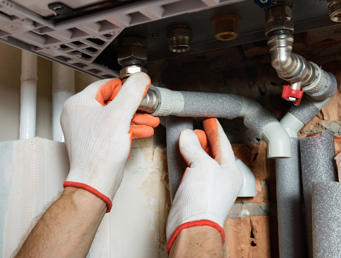 Person in gloves tightening a pipe fitting under a sink, working on plumbing installation with insulated pipes.