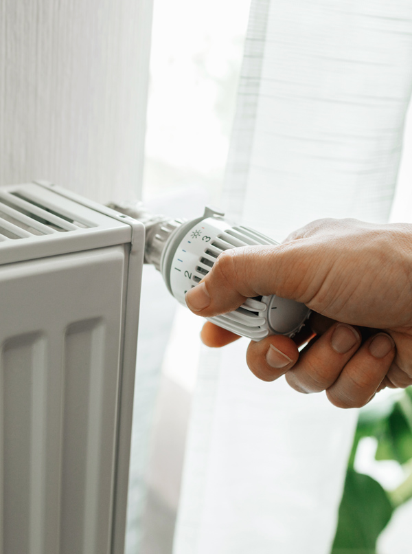 Hand adjusting the thermostat knob on a modern heating radiator, with light filtering through a curtain. Home heating control.