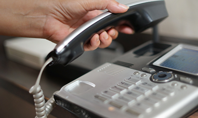 A hand is lifting the receiver of a traditional telephone, with a close-up view of the keypad and screen visible in the background, showcasing an office communication device.