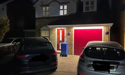 A suburban home with a red garage door illuminated at night, featuring two parked cars in the driveway and a blue recycling bin.