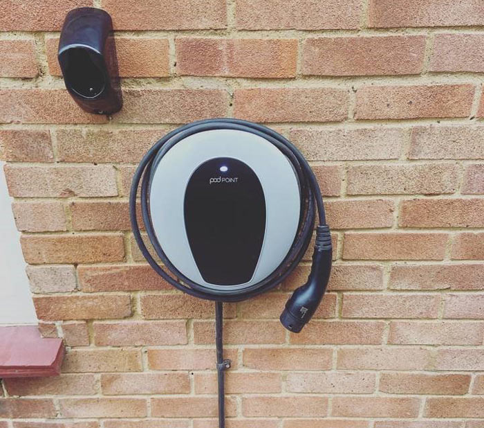 Wall-mounted electric vehicle charger with a coiled cable against a brick wall, showcasing a modern design ideal for home charging solutions.