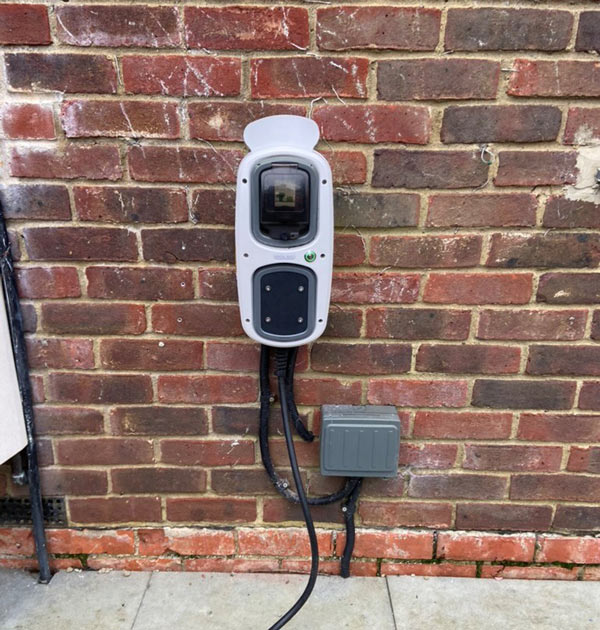 Electric vehicle charging station mounted on a brick wall, featuring a digital display and cables leading down to a gray electrical box.