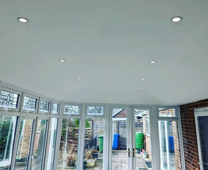 Interior view of a conservatory featuring bright recessed lighting, large windows, and a glimpse of the outdoor area with greenery and storage bins visible outside. Stylish design and natural light create an inviting space.