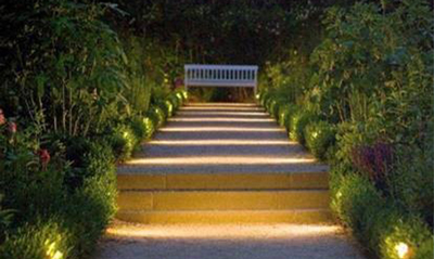 A serene garden pathway illuminated by soft lights, leading to a white bench surrounded by lush greenery.