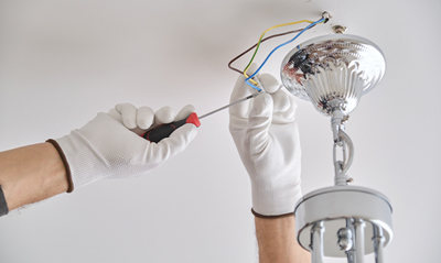 hands wearing gloves using a screwdriver to install a ceiling light fixture, showing electrical wires in a home improvement context